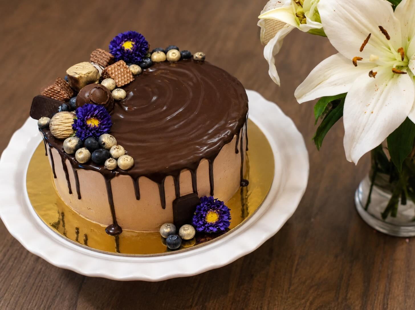 A chocolate cake with a glossy ganache drip, decorated with gilded chocolates, blueberries and violet flowers, beside white lilies
