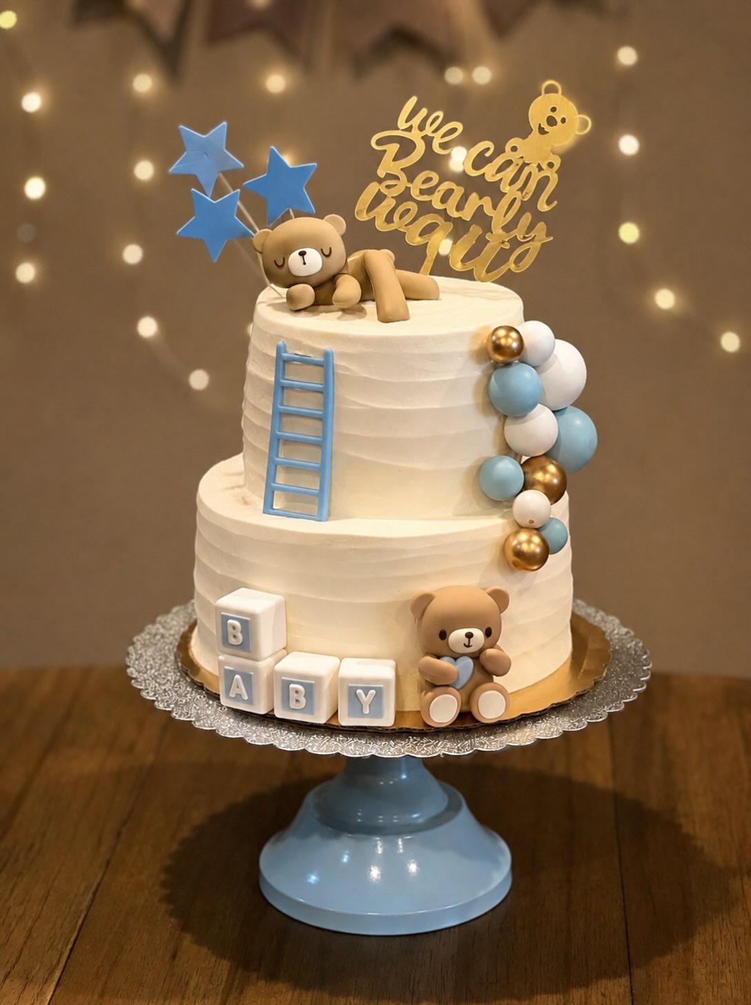A two-tier white baby shower cake with sugar teddy bears, blue alphabet blocks spelling BABY, and blue and gold balloons