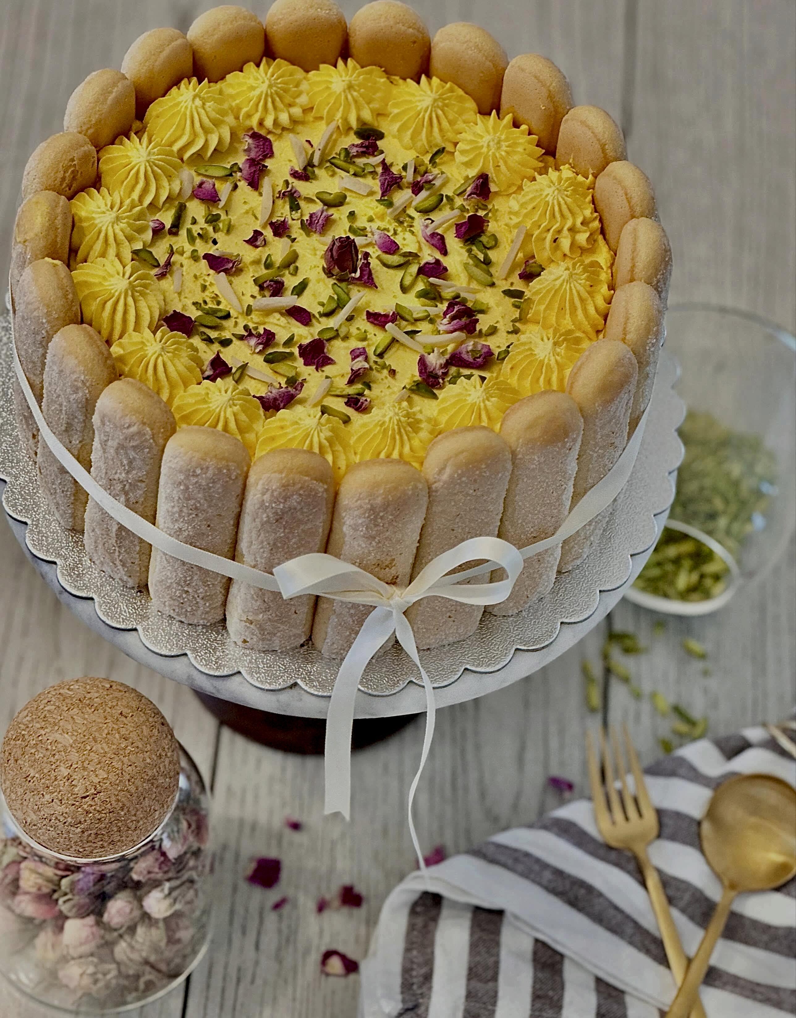 A charlotte cake ringed with ladyfingers and tied with a cream ribbon, its golden piped cream scattered with rose petals and pistachios